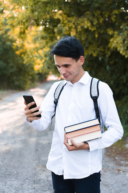 A dark-haired schoolboy, dressed in a white and clean shirt, with a gray briefcase on his shoulders, holds a phone and multi-colored books on an autumn background.