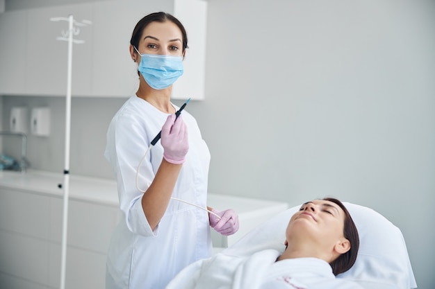 Dark-haired woman doctor in latex gloves and a face mask holding a heated needle electrode