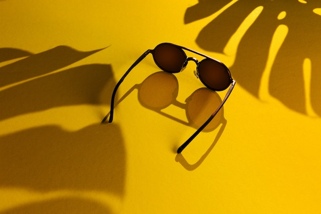 Dark metal sunglasses with shadow of monstera palm on abstract yellow background