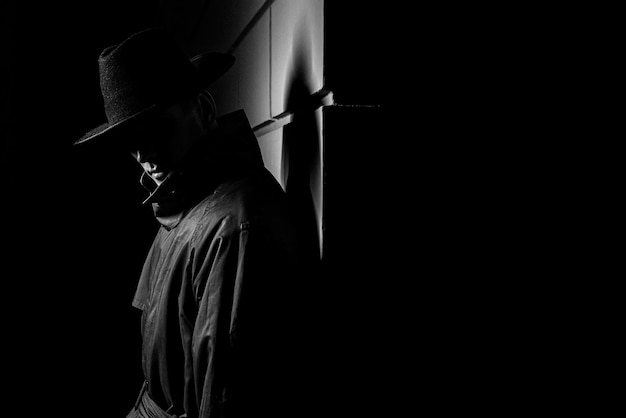 Dark silhouette of a man in a raincoat with a hat at night on the street in a crime Noir style