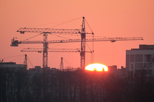 Dark silhouette of tower cranes with big setting sun at high residential apartment buildings construction site at sunset Real estate development