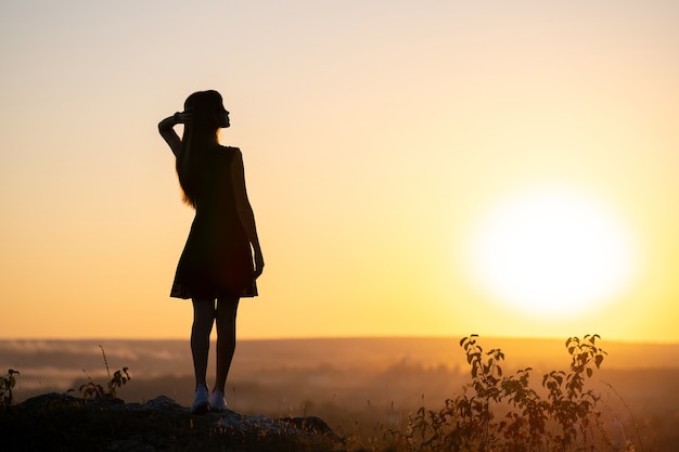 Dark silhouette of a young woman in summer dress standing outdoors enjoying view of nature at sunset.