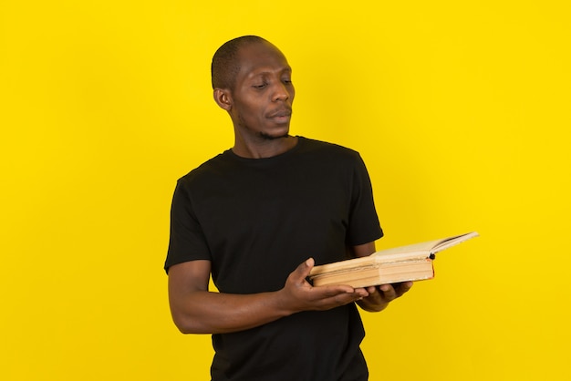 Dark-skinned young man holding book and thinking on yellow wall