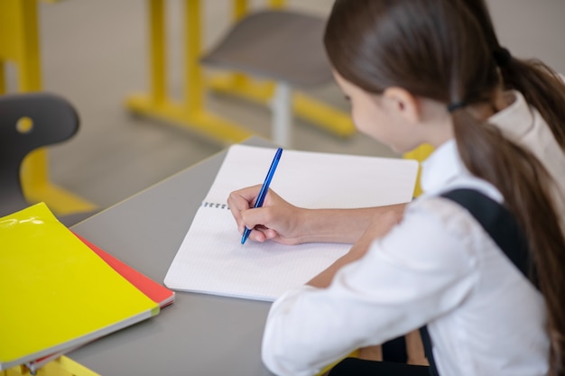 Darkhaired girl in school uniform is diligently writing with pen in notebook sitting at school desk