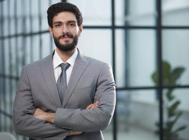 A darkhaired man in an office in a suit stands with crossed arms in front of him