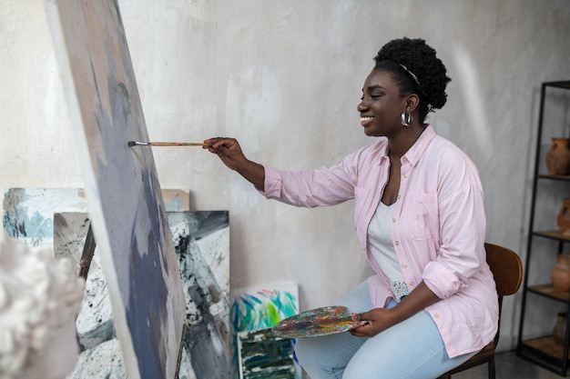 A darkskinned woman sitting near the easel and painting