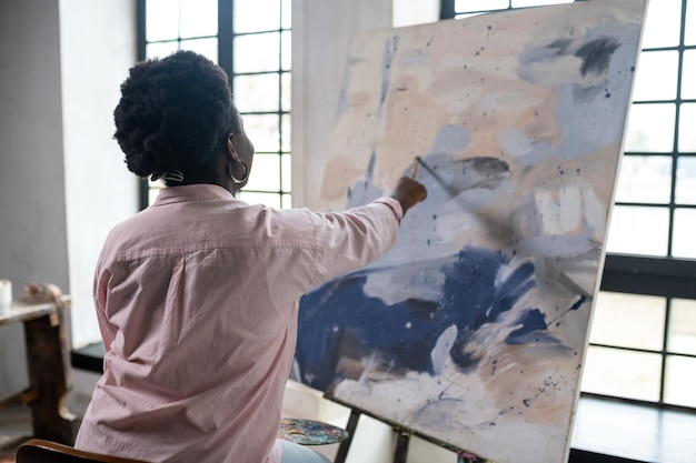 A darkskinned woman sitting near the easel and painting