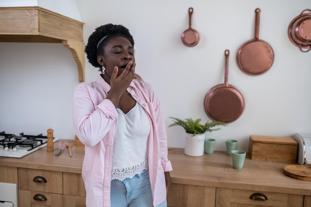 A darkskinned woman standing in the kitchen and yawning