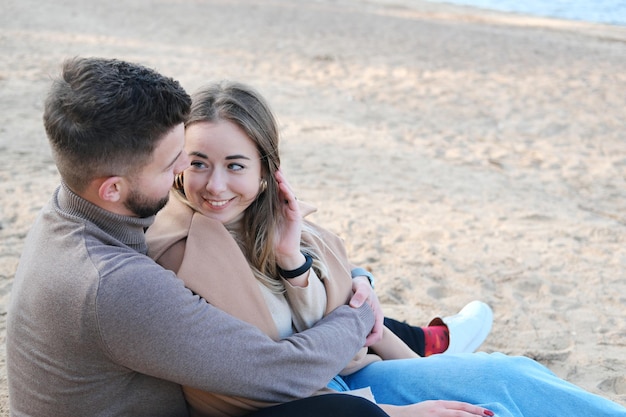 Date on the beach in the cold season The guy hugs the girl from behind by the shoulders and tries to warm her