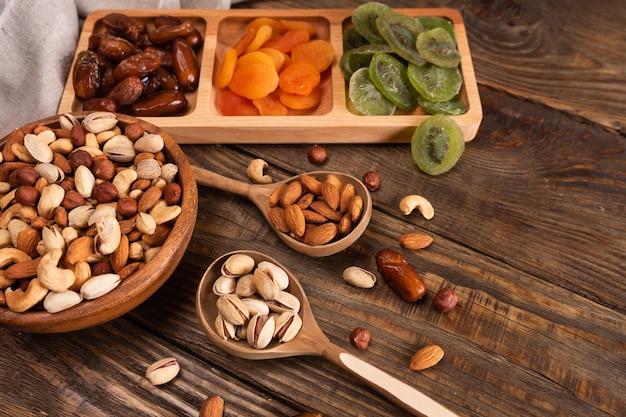 Dates, dried apricots and kiwis in a Compartmental dish and assortment of nuts in wooden bowl on a dark wooden table.