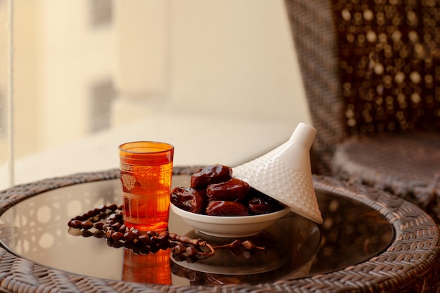 Dates in a glass plate with a lid, an Oriental drink and a rosary on a glass table