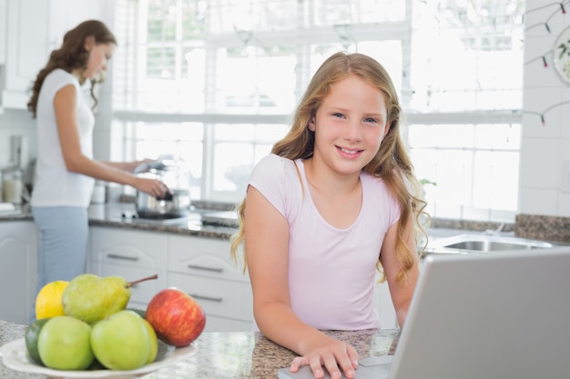 Daughter using laptop with mother cooking food in background at kitchen