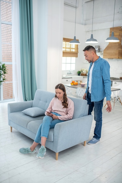 Daughter with a smartphone in her hands in headphones sitting on a sofa listening to music but not listening to her father