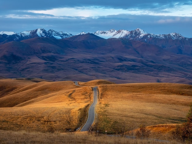 Dawn road stretching into the distance through the picturesque autumn hills Winding asphalted clean road stretches into the distance to the snowcapped mountains