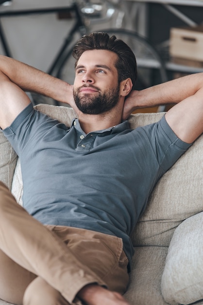 Day dreaming handsome. Handsome young man keeping hands behind his head and looking away while sitting on the couch at home