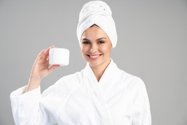 Dazzling young woman preparing to applying moisturizing cream on her face. Photo of woman in white bathrobe and towel on grey background. Skin care concept