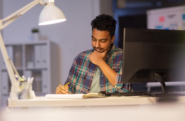 deadline technology and people concept creative man with computer working at night office and making notes in notebook