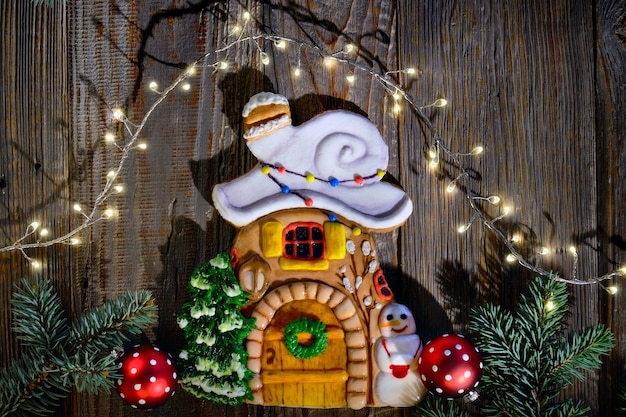 Decorated gingerbread house with snow on the roof, Xmas tree and snowman. Christmas flat lay with fir twigs, red baubles and stars confetti. Dark wooden planks, top view of rustic wood table.