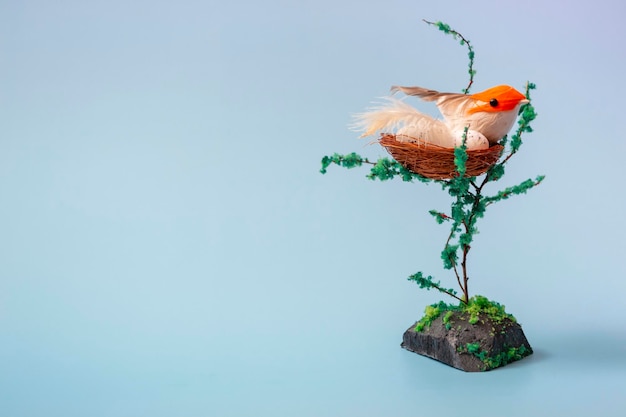 On a decorative tree, a nest with eggs and a small yellow bird on a blue background close up with copy space