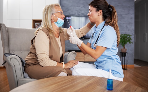 Dedicated nurse with sitting at home with senior woman and taking swab from her nose to test her on coronavirus.