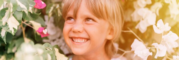 Defocused blurred portrait of face candid little happy smiling five year old blonde kid boy with green eyes in pink and white flower plants in nature children have fun summer holidays banner flare