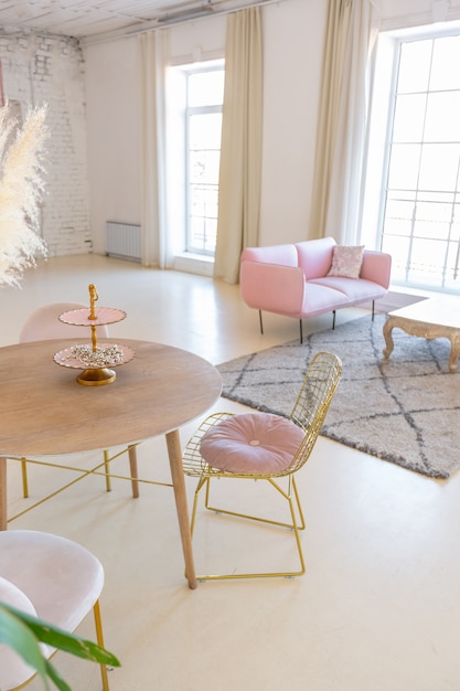 Delicate and cozy light interior of the living room with modern stylish furniture of pastel pink color and white walls with stucco moldings in daylight