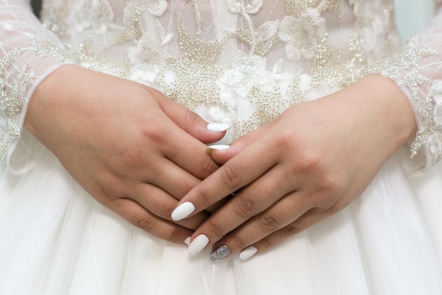 Delicate hands of the bride with a white manicure on the background of the wedding dress