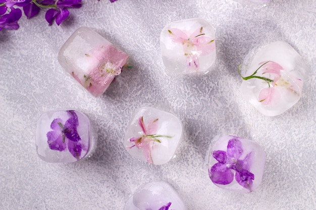 Delicate pink and purple flowers frozen in ice cubes on a light gray background. Top view