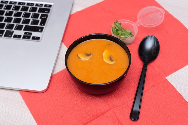 Delicious asian tom yum soup in takeout packaging with spoon and laptop on office table Selective focus Asian delivery food concept Healthy eating