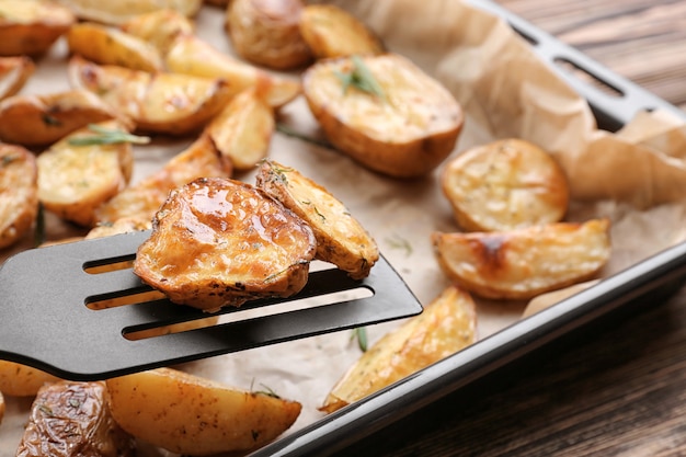 Delicious baked potatoes with rosemary on spatula above baking tray, closeup