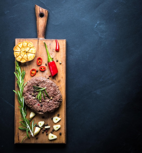 Delicious beef burger steak with spices and herbs on cutting board and slate background top view