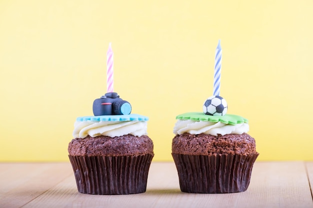 Delicious birthday cupcakes with icons of ball and camera on it and candles on wooden desk and yellow background