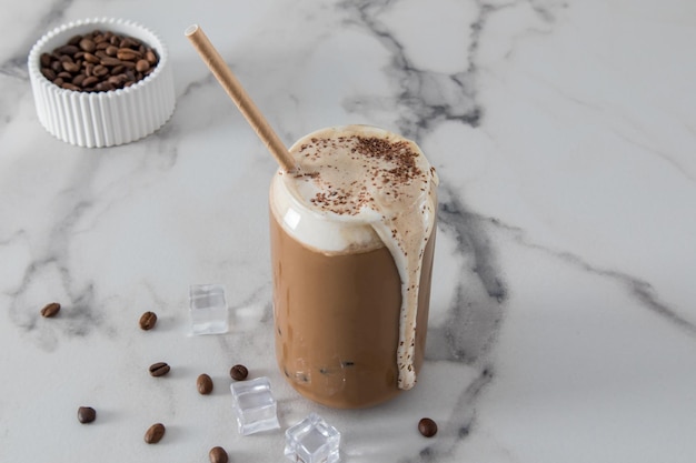 Delicious coffee latte in a modern stack in the shape of a can with foam flowing down the wall of the glass front view marble white background