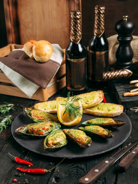 Delicious cooked mussels with sauce, lemon and garlic bread and butter on a black plate on the dark wooden background