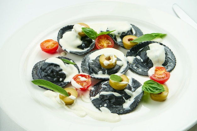 Delicious cuttlefish ink ravioli stuffed with shrimps and scallops, served with white sauce, cherry tomatoes and olives in a white plate on white tablecloth