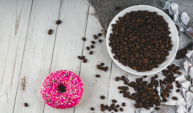 A delicious donut near a small white plate with coffee beans The coffee beans are scattered on the table for decoration Close up