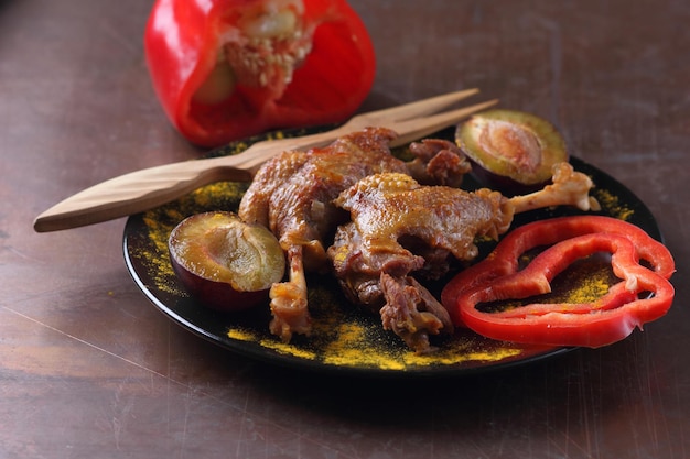 Delicious duck legs with golden crust closeup Two baked duck legs with red bell pepper plums and turmeric on a black plate on a dark background