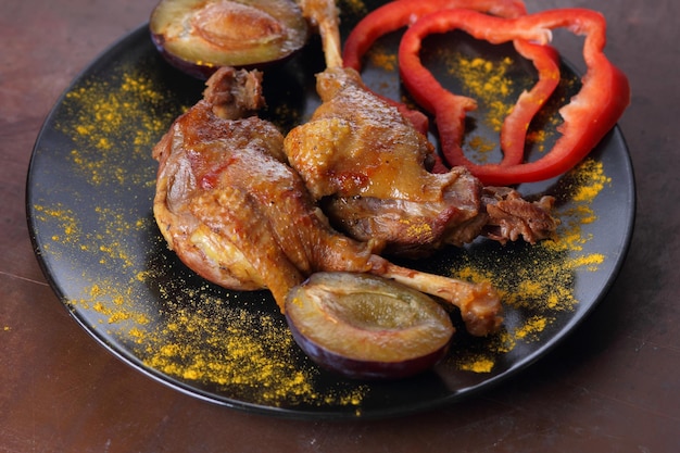 Delicious duck legs with golden crust closeup Two baked duck legs with red bell pepper plums and turmeric on a black plate on a dark background