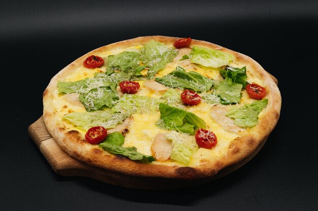 Delicious fresh cesar pizza with chicken and vegetables on wooden board on black surface