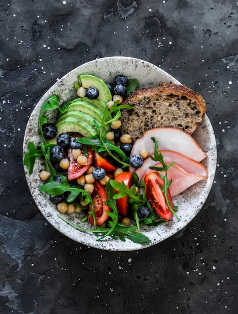 Delicious healthy breakfast snack ham and avocado arugula chickpeas tomatoes blueberry salad on a dark background top view