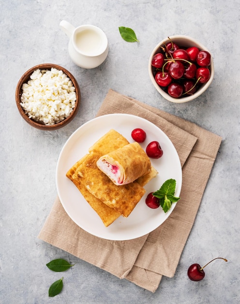 Delicious homemade pancakes or crepes with cottage cheese and fresh cherries on a white plate with sour cream on a blue background Top view