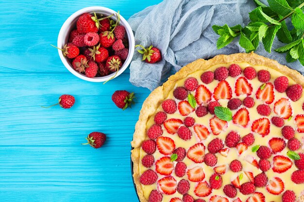 Delicious homemade tart with strawberries and raspberries garnished mint leaves on blue wooden background. Top view