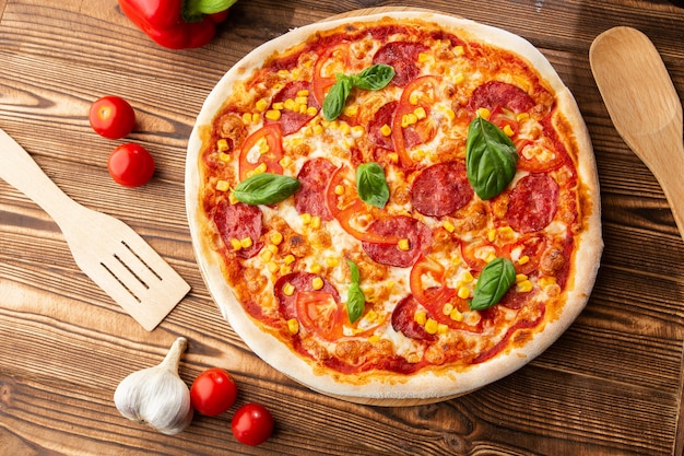 Delicious hot homemade pizza on the wooden table. Pizza - Fresh homemade pizza with pepperoni, cheese and tomato sauce, tomatoes, sweet corn and basil with copy space.