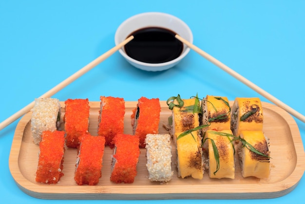 Delicious Japanese rolls with salmon avocado cucumber cheese on wooden plate with wood sticks and soy sauce with selective focus on blue background Asian food concept Healthy eating