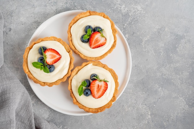 Delicious mini tarts with fresh berries and custard on a white plate on a gray concrete background