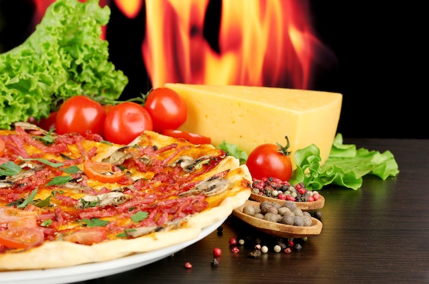 Delicious pizza salami tomatoes and spices on wooden table on flame background