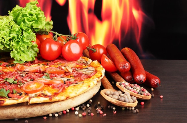 Delicious pizza salami tomatoes and spices on wooden table on flame background