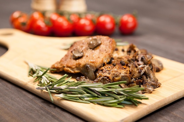 Delicious pork steak on wooden board next to a branch of oregano and cherry tomatoes. Two shakers with salt and pepper are blurred in the background