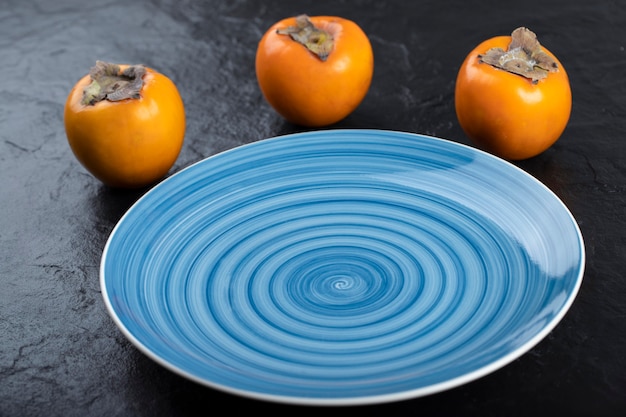 Delicious ripe persimmon fruits and empty blue plate on black surface.