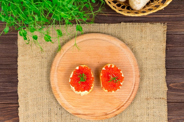 Delicious sandwiches with red caviar and butter on a wooden board against a background of herbs and mushrooms View from above